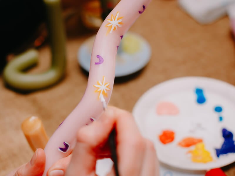 A close up of a person holding a bendy pink candle and painting small stars and moons on it